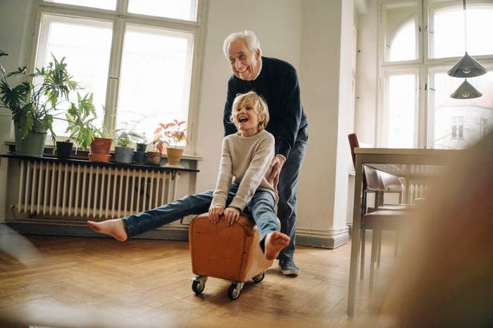 An older man pushes a joyful child seated on a small wheeled suitcase in a bright, plant-filled room with large windows