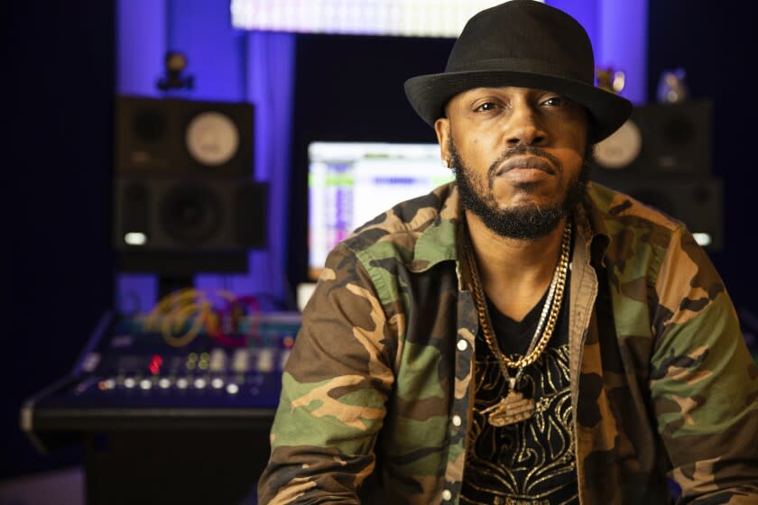 A man in a black hat, gold chains and camo jacket poses for a portrait