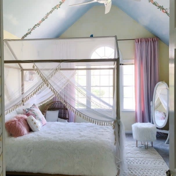 a reviewer photo of the canopy bed with sheer drapery in a bedroom with a vaulted ceiling, adjacent to a wicker chair and a vanity mirror