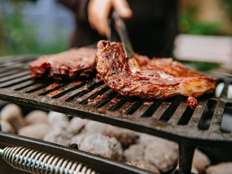 a person grilling steaks with metal tongs on a charcoal grill