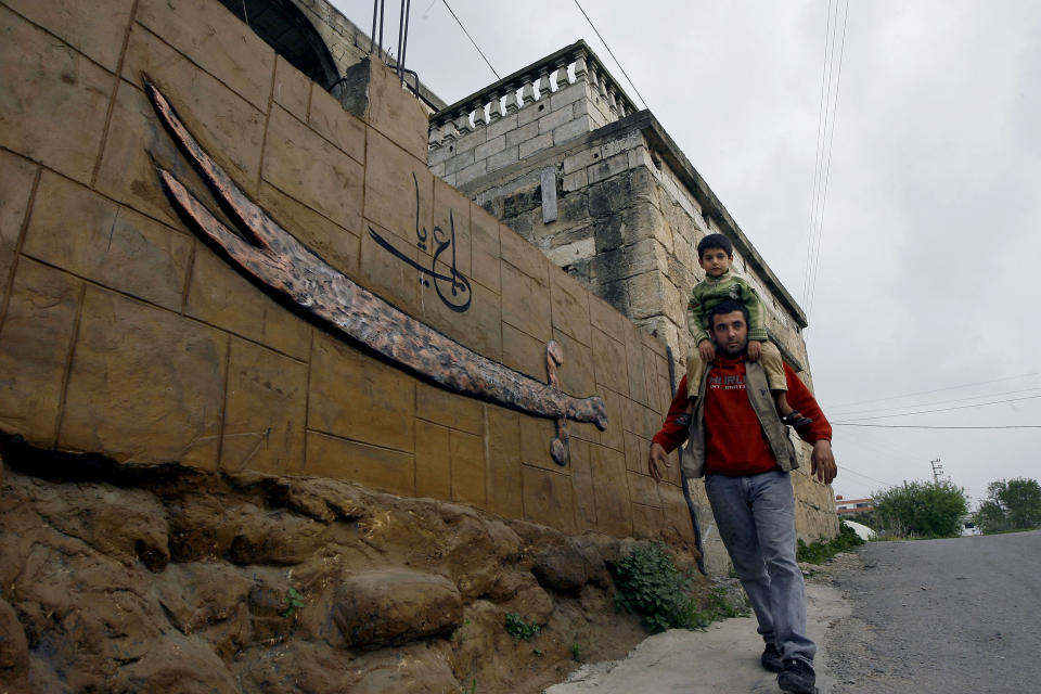 In this picture taken on Monday, Feb. 24, 2014, a Shiite man carrying his son on his shoulders passes in front of suicide bomber Nidal al-Mughayar's family home in the southern village of Bisariyeh, Lebanon. Nidal al-Mughayar renewed his travel document last year and told his family he will be leaving Lebanon to settle in Venezuela where there are more opportunities but it turned out later that the young Palestinian man called his family from Syria and it was only then that they knew he has joined jihadis fighting to overthrow President Bashar Assad’s government. (AP Photo/Mohammed Zaatari)