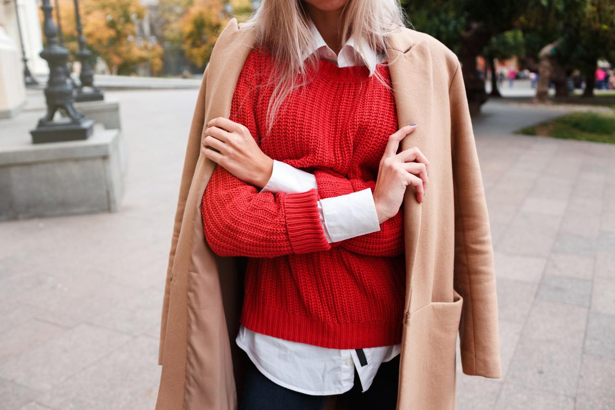 Woman wearing tan coat, red sweater, and white button-up blouse.