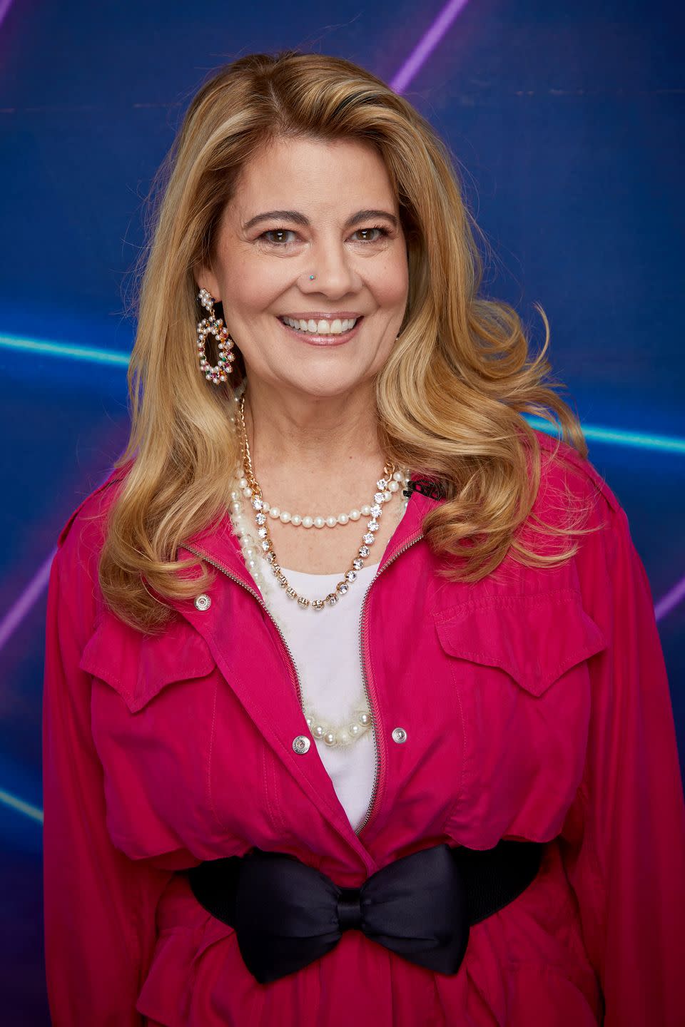 lisa whelchel smiles at the camera, she wears a pink jacket with a bow around the waist and a white shirt as well as several necklaces and large pearl earrings