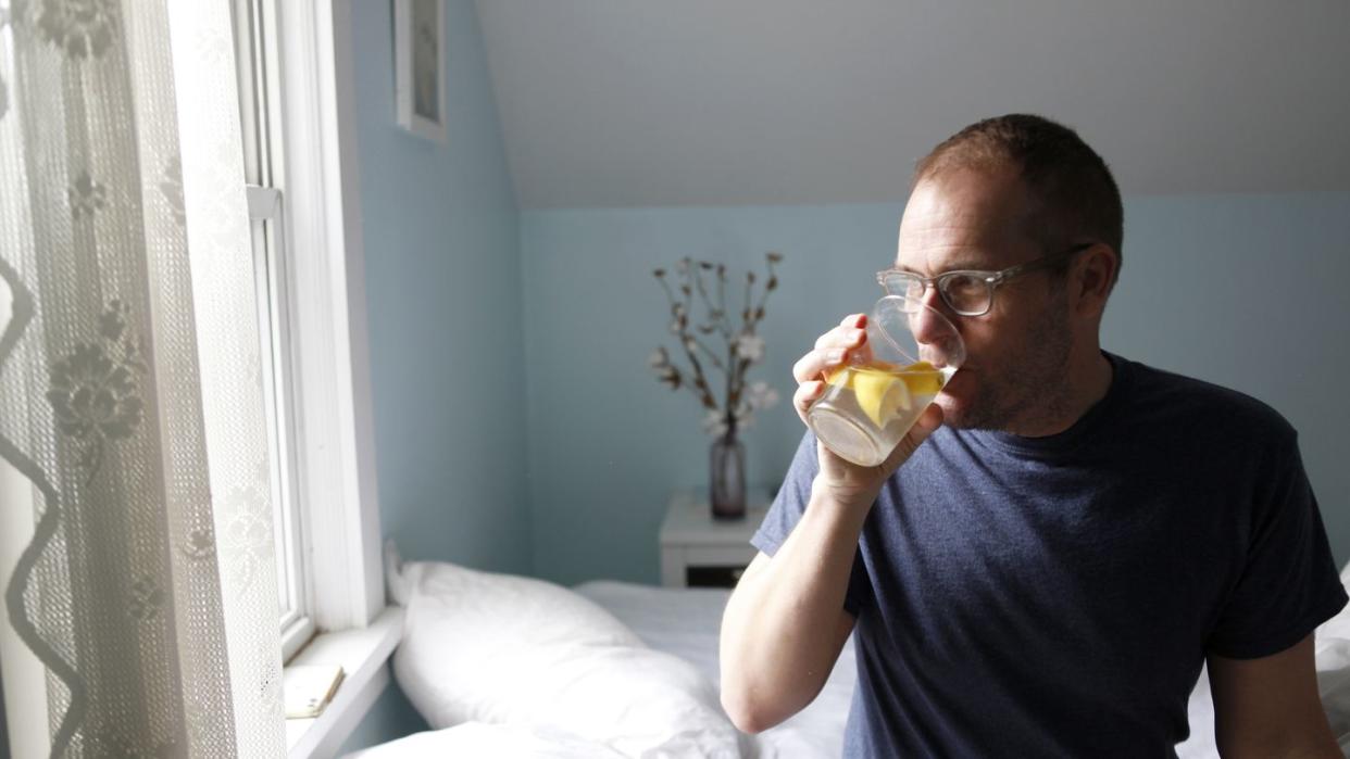 man sitting on side of bed, drinking water with lemon, side view