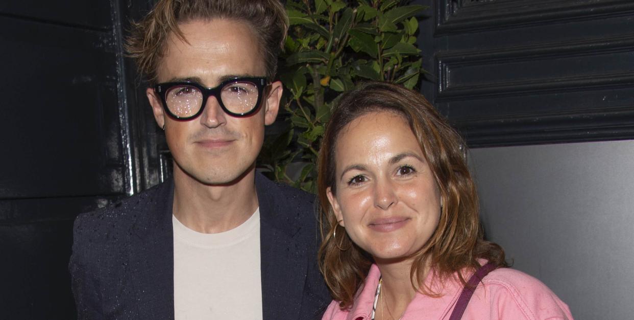 tom fletcher, a man wearing a white shirt and navy suit with thick black glasses, and giovanna fletcher, a woman with short brown hair wearing a pink demin jacket, pose and smile together in front of a leafy plant