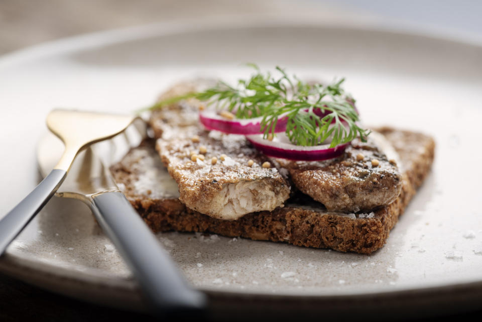 A plate with a slice of rye bread topped with a fish fillet, onion, and herbs, with cutlery on the side