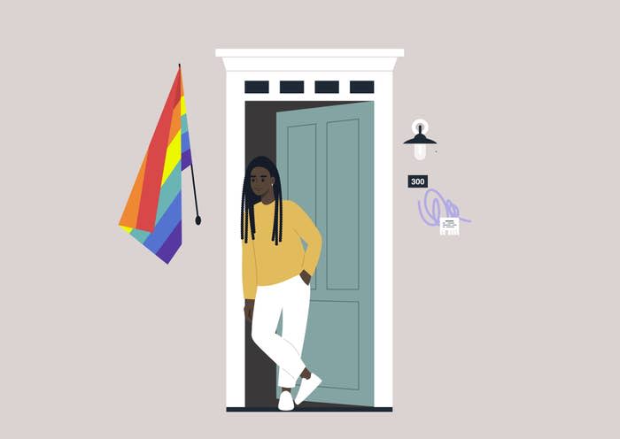 Person with long braids, in a sweater and pants, stands at an open door. A rainbow pride flag is hung beside them. House number 300 is visible