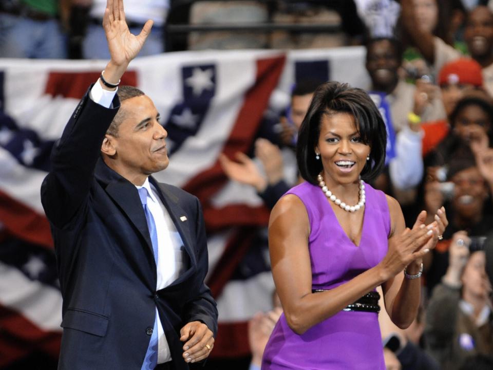 Barack Obama and Michelle Obama wave and clap at a Chicago campaign. Michelle Obama is wearing a purple cocktail dress and pearl necklace.