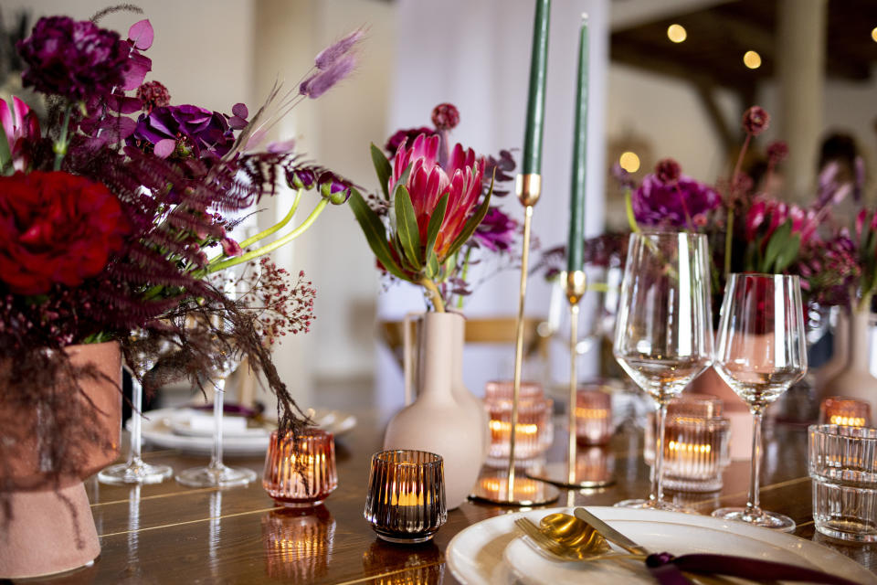 Elegant table setting featuring a variety of flowers in vases, candles, glassware, and gold-accented cutlery on a wooden table