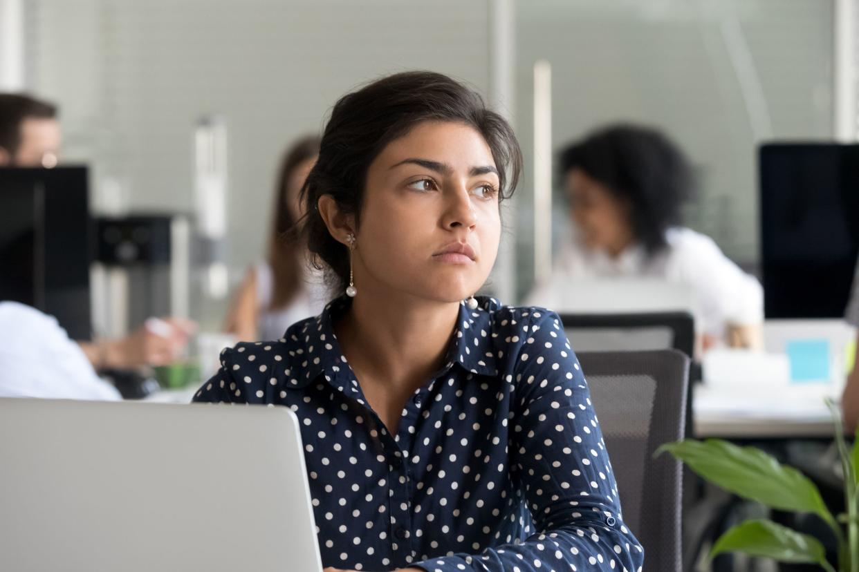 Thoughtful young woman sitting at a desk in an office, facing the camera, looking off in the distance towards the right, several work colleagues at desks blurred in the background