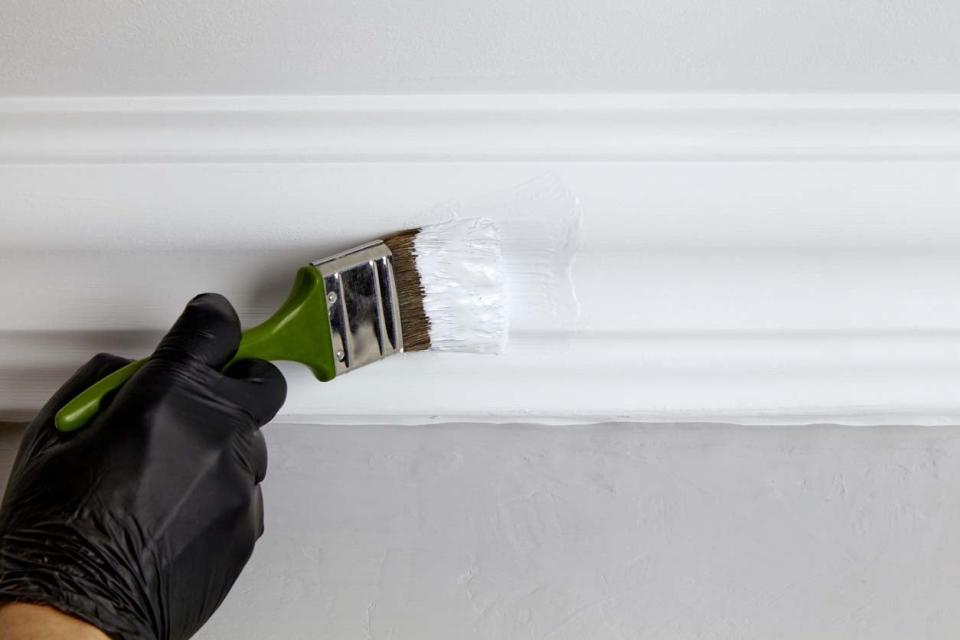 A close up of a person's gloved hand using a paint brush to paint trim white. 