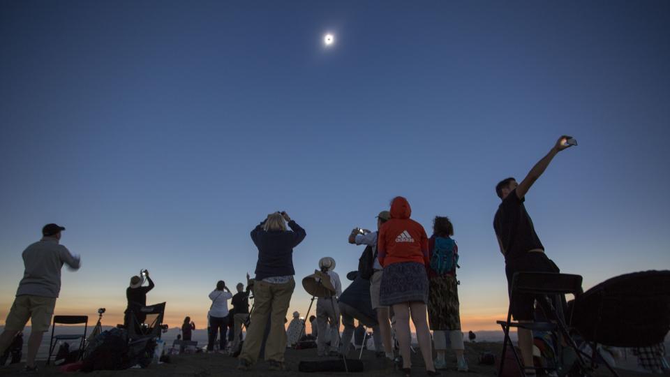 people on a hillside look upward at the sun, some holding cameras or telescopes