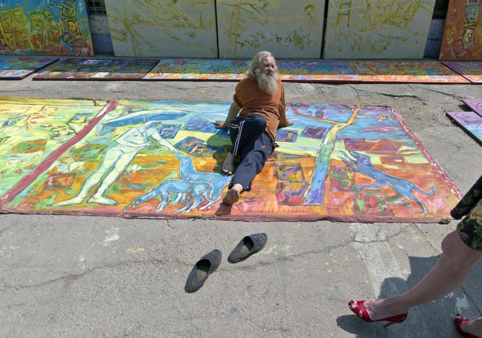 In this photo taken on April 4, 2013, Romanian artist Vasile Muresan, known as Murivale sits on one of his paintings in a parking lot, in Bucharest, Romania. The white-bearded painter can often be seen sitting on his colorful canvasses which he displays in the street followed by the street dogs which are his companions and also inspire his work. The 56-year-old, whose home city is Bistrita_the Transylvanian town associated with the legendary Count Dracula_ has been painting with passion since he was a teen, producing vivid works of Monaco, the streets of Paris, the hurly burly of the Romanian capital and huge colorful more abstract canvasses. (AP Photo/Vadim Ghirda)