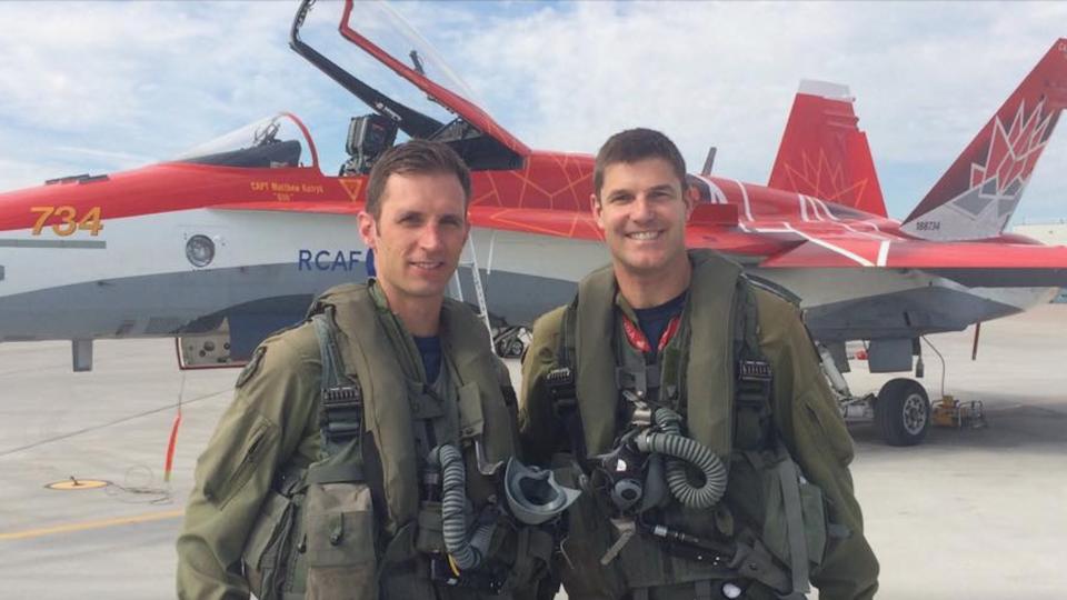 two men in flight suits stand in front of a jet painted red and white