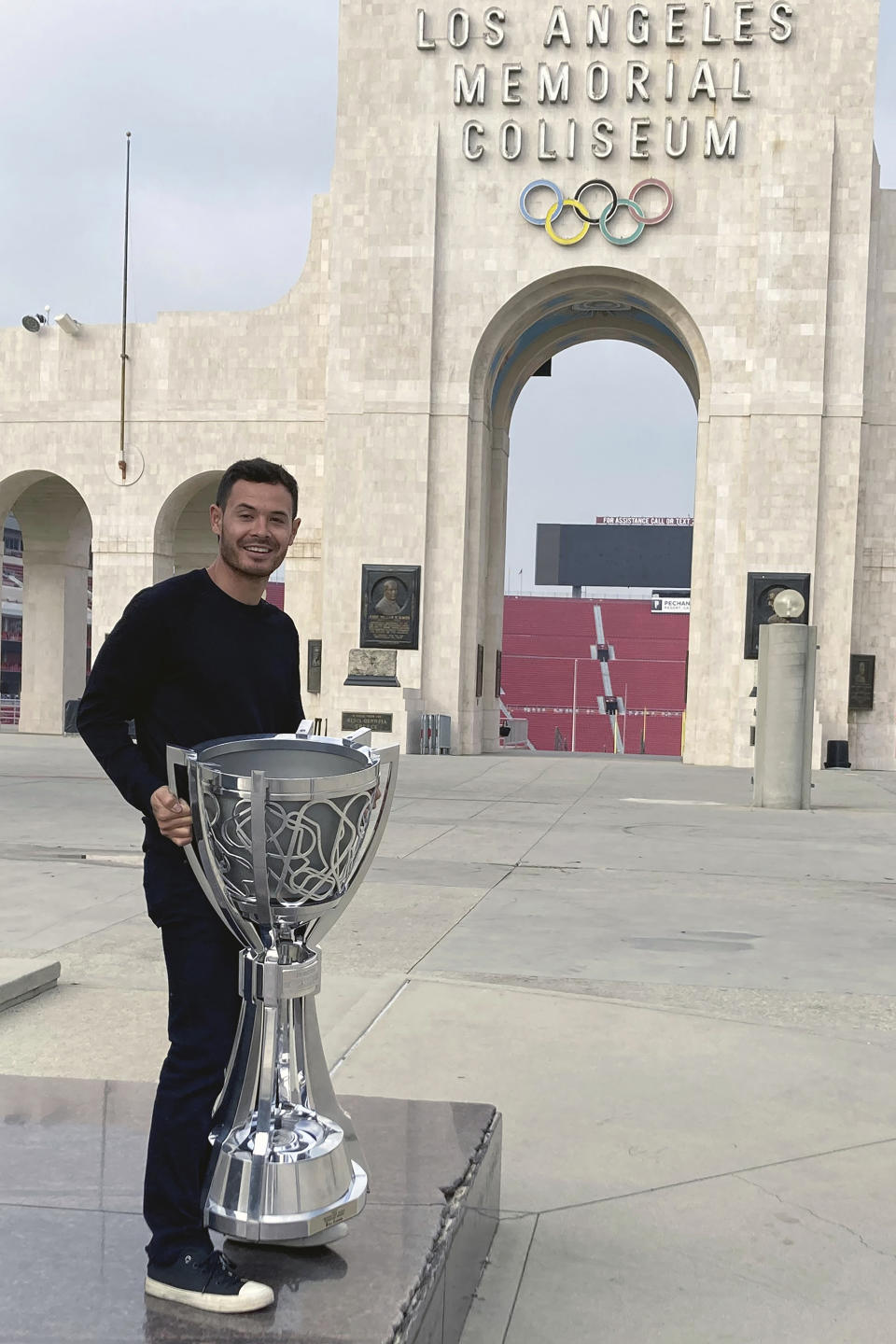 NASCAR driver Kyle Larson poses with the NASCAR Cup Series championship trophy in front of the Los Angeles Memorial Coliseum, Tuesday, Nov. 9, 2021, two days after winning the series championship in Avondale, Ariz. Larson toured Los Angeles Memorial Coliseum on Tuesday, the first driver to get an up-close look at what NASCAR plans when it shifts to the season-opening exhibition Busch Clash Feb. 6 on a purpose-built track inside the Coliseum. Larson, as the champion, is the only driver locked into the field so far.(AP Photo/Jenna Fryer)