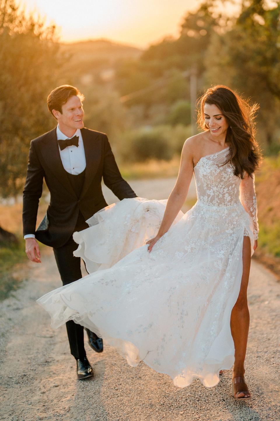 A groom helps his bride carry her wedding dress train up a hill.