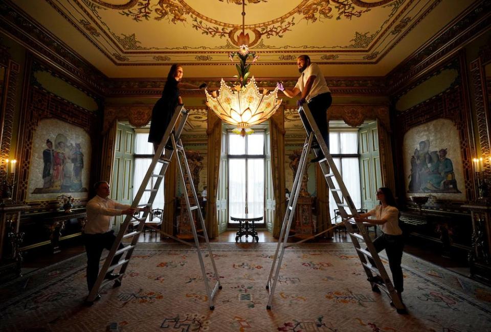 Dois funcionários do Royal Collection Trust ficam em escadas um de frente para o outro enquanto cuidam de um grande e brilhante lustre de vidro em forma de lótus na Ala Leste do Palácio de Buckingham, em Londres, em 8 de junho de 2024, após sua reforma.  Atrás deles fica a varanda usada pelos membros da família real para aparições públicas.