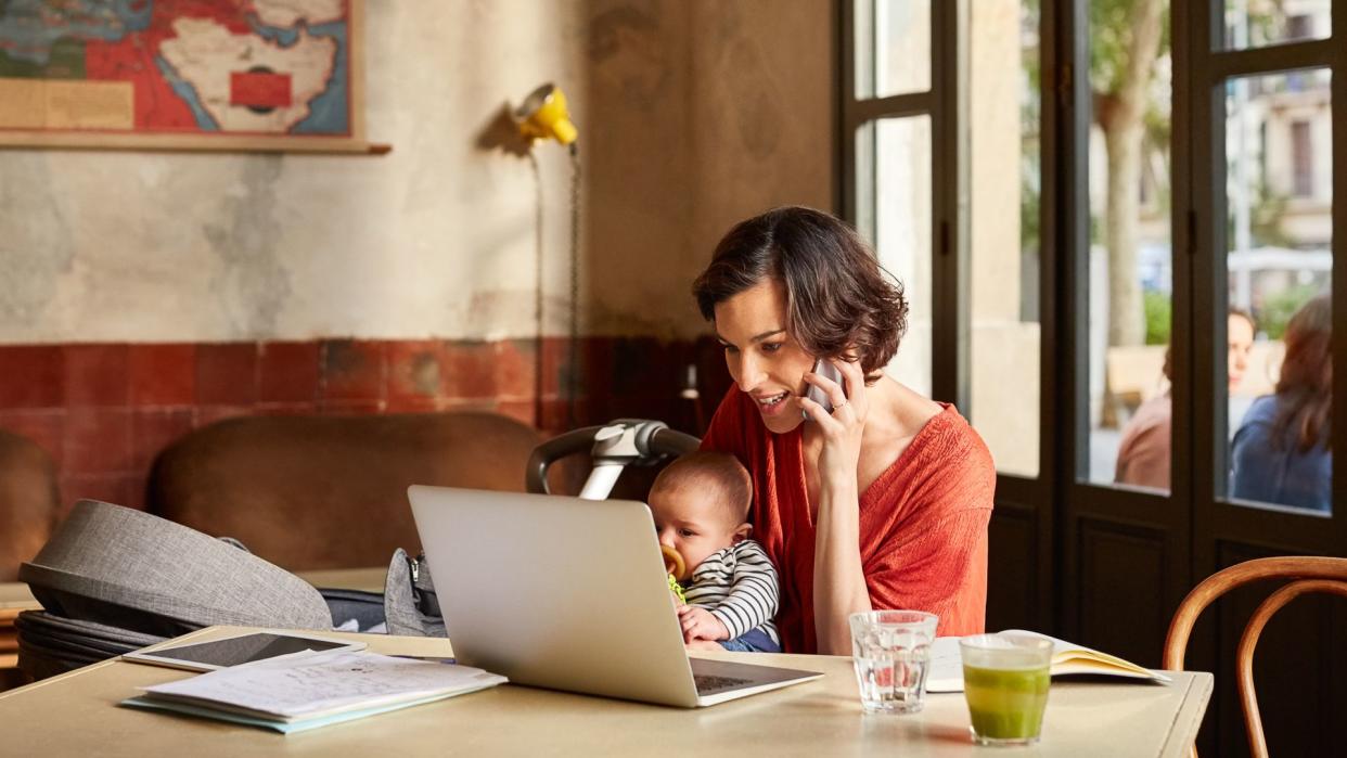 Mother carrying baby using phone while looking in laptop at table in restaurant.