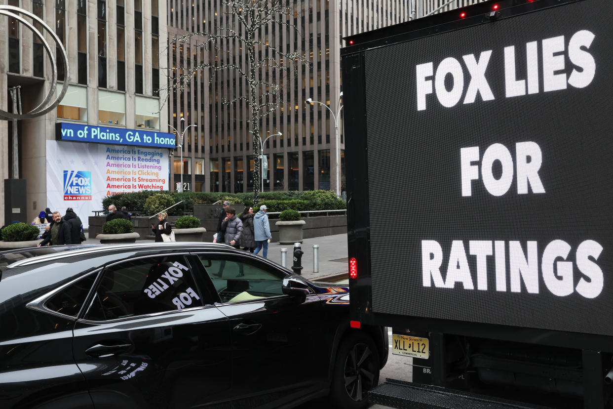 A video screen on the back of a truck, in front of an array of office buildings, displays a message: Fox Lies for Ratings.