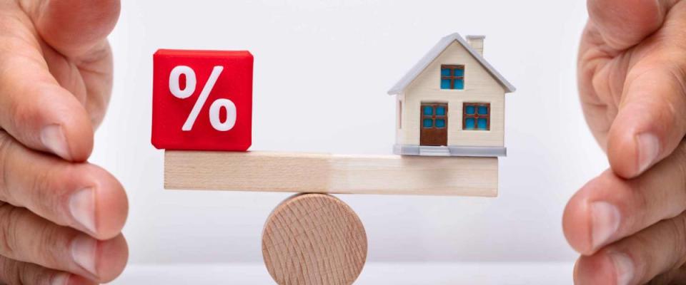 Businessperson's Hand Protecting Balance Between Percentage Red Cubic Block And House Model On Wooden Seesaw