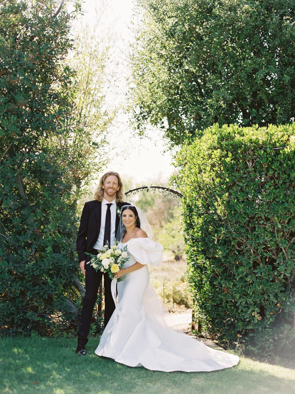 A bride and groom pose in front of two green trees.