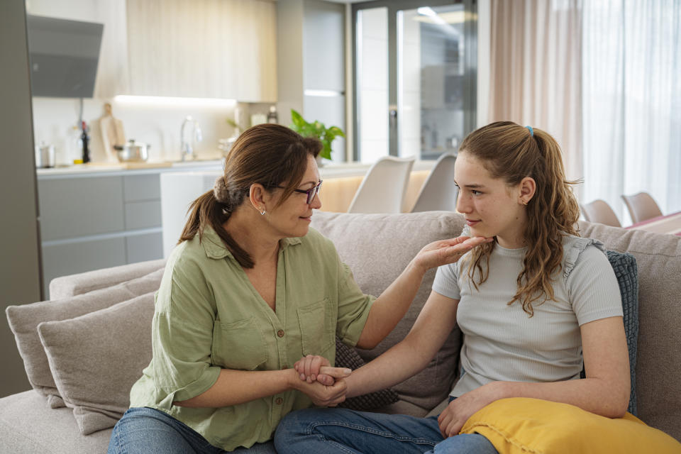 An older woman in a green shirt sits on a couch, holding the hand of a younger girl in a gray shirt, comforting her. They are in a modern living room