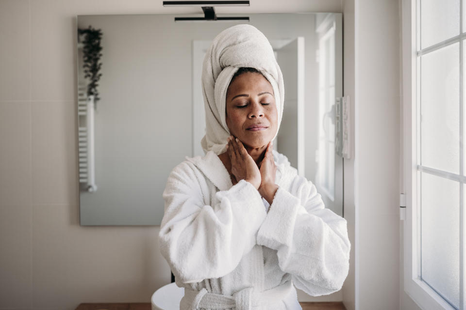 Woman with a towel around her head, wearing a bathrobe and standing in front of a mirror with her eyes closed and her hands on her neck