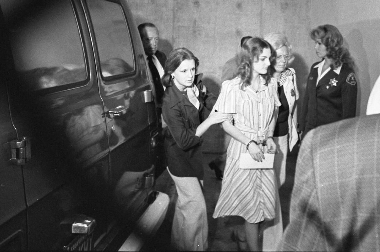 Patricia Hearst, wearing a striped dress and in handcuffs, is escorted by two women at the inmate court entrance