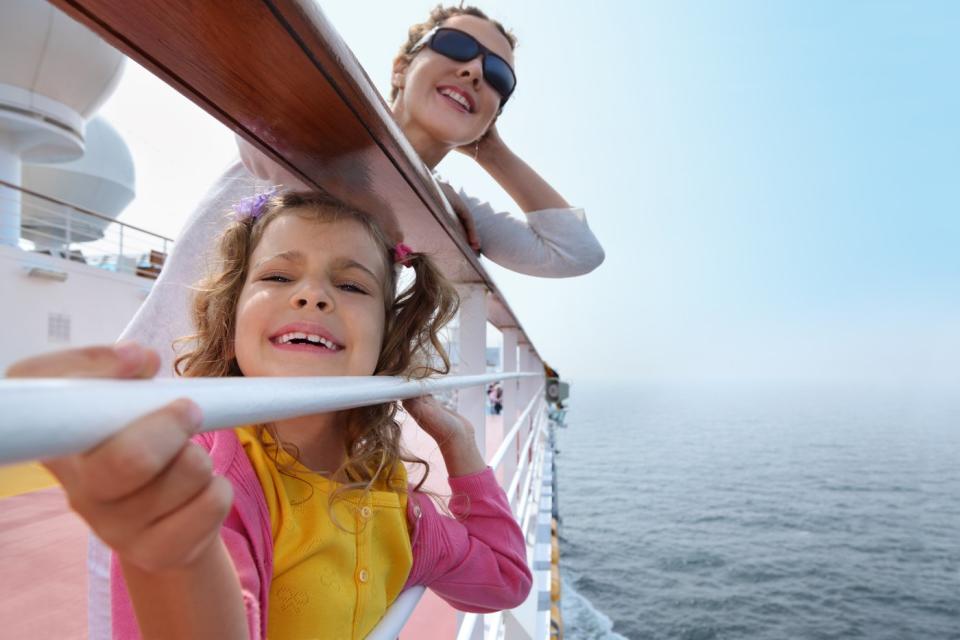 A parent and child smile while holding onto a railing on the deck of a cruise ship.