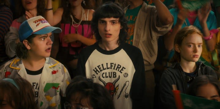Gaten Matarazzo, Finn Wolfhard, and Sadie Sink stand in a crowd during a high-school rally in a scene from Stranger Things 4.