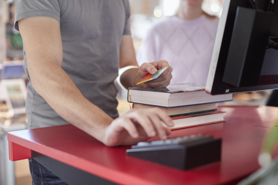 A person pays for a stack of books at a store counter. Another customer stands nearby