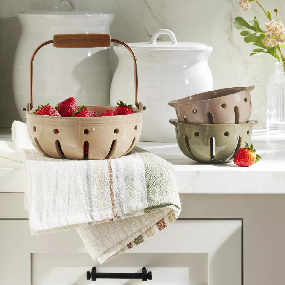 The taupe bowl featuring a round shape with holes and space below to let the water drain out after washing, and a metal holder with a wooden handle