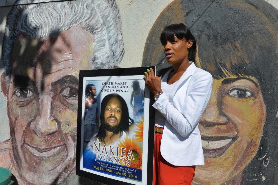 In this April 16, 2014, photo, Shackelia Jackson-Thomas holds a poster showing her brother, Nakiea Jackson, just outside his small “cookshop” in the Orange Villa section of Jamaica’s capital of Kingston. Earlier this year, the 27-year-old cook was shot dead by a policeman as pots of curried goat simmered on the stove of his kitchen, the exterior of which is decorated with murals of past and present political leaders. The patrolman who shot him is one of 27 police officers now facing charges of murder brought by an investigative commission probing allegations against police and soldiers. Jamaica’s security forces have long been accused of indiscriminate shootings and unlawful killings. (AP Photo/David McFadden)