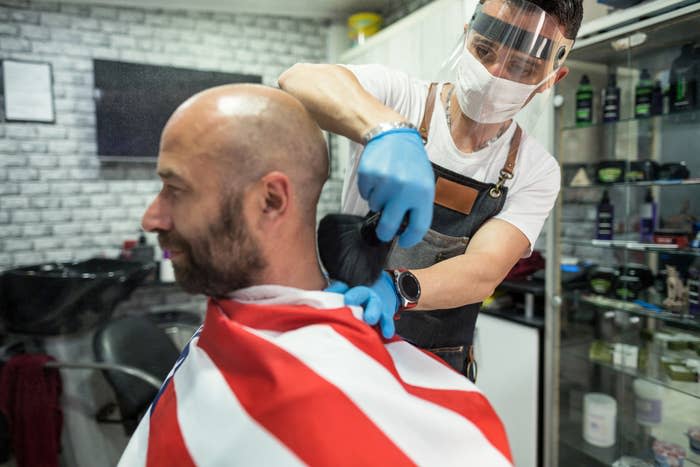 Barber wearing a surgical mask, gloves, and a face shield brushes hair off a customer's neck