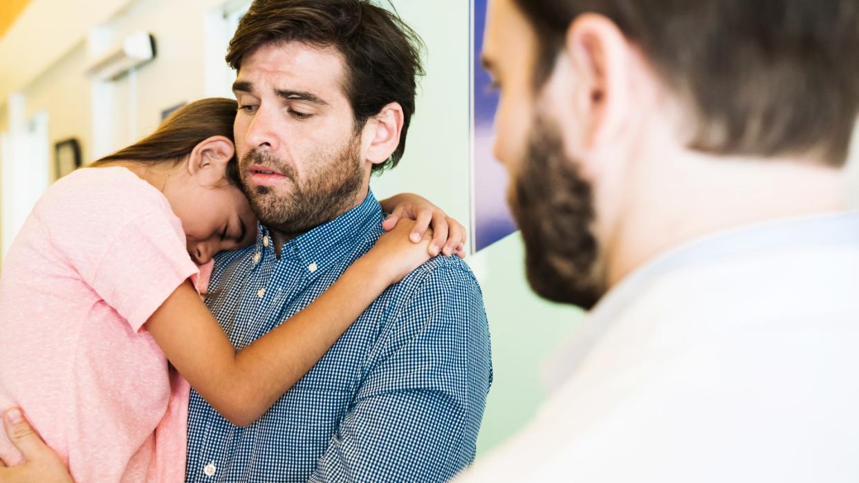  Father carries his daughter who buries her head into his shoulder in front of a doctor in the foreground. 