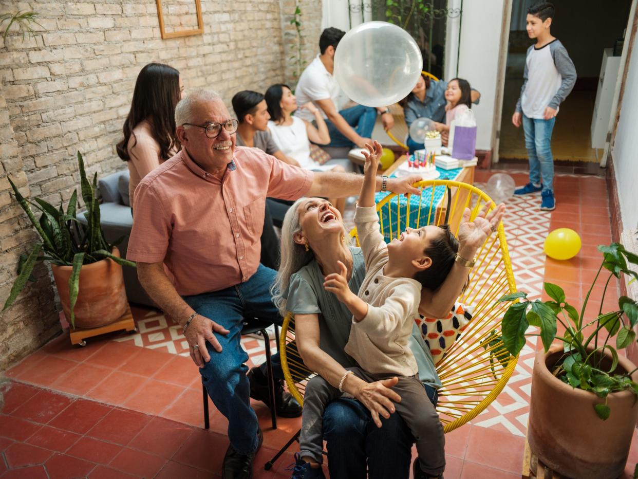 Happy mexican parents sitting with their grandson on chairs and playing with a balloon during a birthday celebration at home.