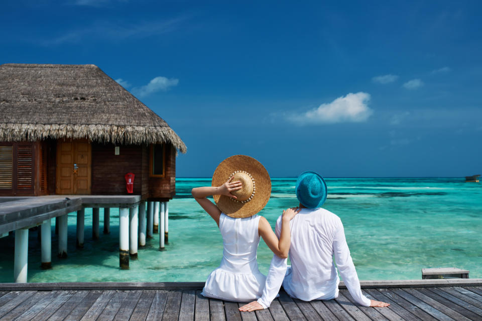 A couple sitting on a wooden walkway looks out over the ocean, with thatch-roof villa to the left