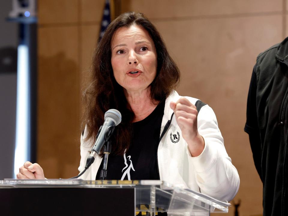 Fran Drescher speaks at a microphone on a podium, wearing a white jacket, with her fist raised.