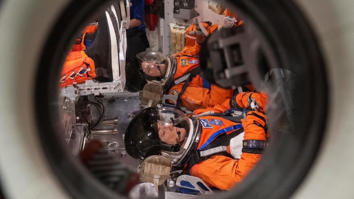  A look through a circular spacecraft window to see two astronauts in spacesuits lying on their backs . 