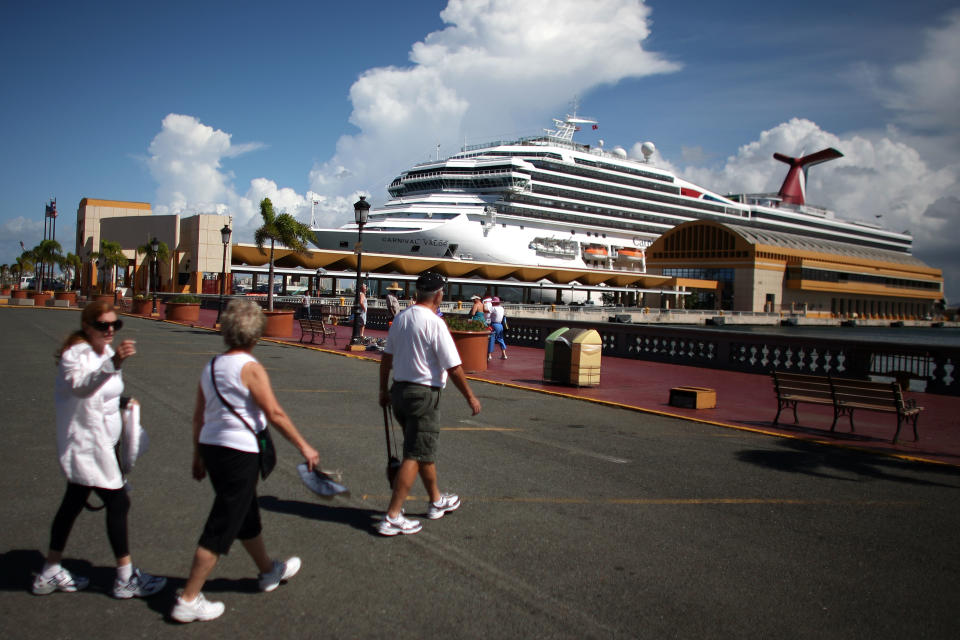 In this Oct. 2, 2012 photo, tourists walk through a parking lot in front of a pier where a cruise ship is docked in Old San Juan, Puerto Rico. Trade groups say the flourishing cruise ship industry injects about $2 billion a year into the economies of the Caribbean, the world’s No. 1 cruise destination, but critics complain that it produces relatively little local revenue because so many passengers dine, shop and purchase heavily marked-up shore excursions on the boats or splurge at international chain shops on the piers. (AP Photo/Ricardo Arduengo)