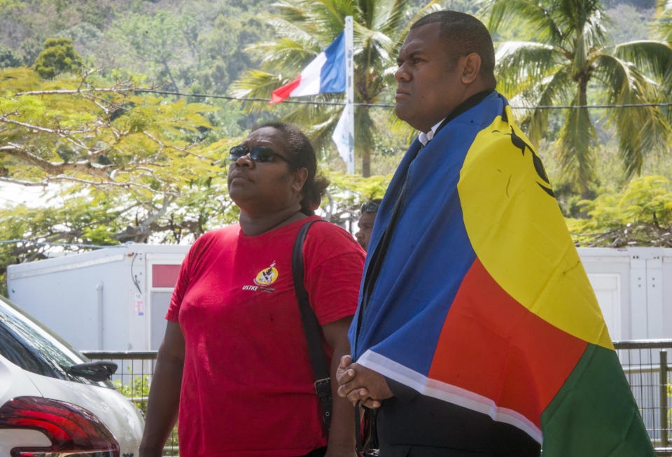 A man, right, drapes the flag of New Caledonia on his shoulders as the French flag flies in the background as he and a woman line up to cast their votes at a polling station in Noumea, New Caledonia, as part of an independence referendum, Sunday, Nov. 4, 2018. Voters in New Caledonia are deciding whether the French territory in the South Pacific should break free from the European country that claimed it in the mid-19th century. (AP Photo/Mathurin Derel)