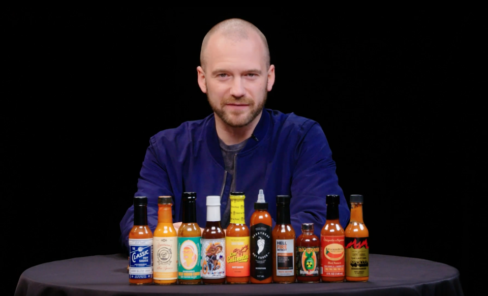 Sean Evans sits behind a table displaying various hot sauce bottles