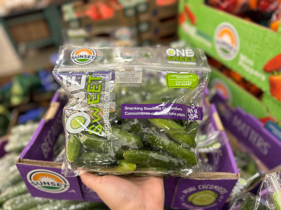 A hand holds a clear plastic bag of snacking cucumbers in front of a purple cardboard box at Costco