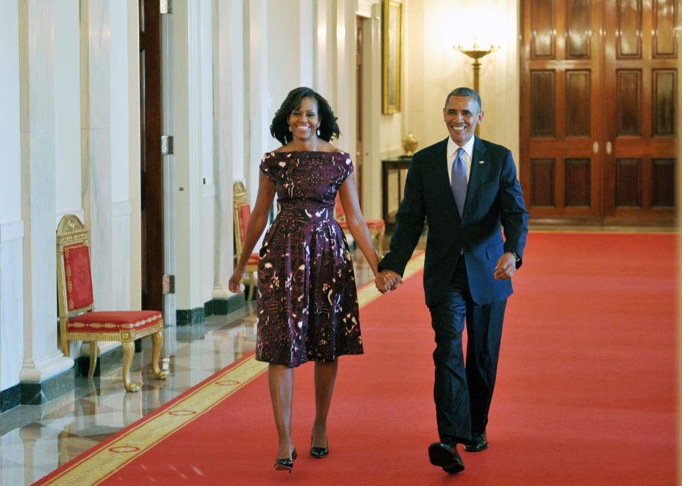 Barack Obama and Michelle Obama walk hand-in-hand down a red-carpeted hallway, Michelle in an elegant knee-length dress, and Barack in a suit and tie