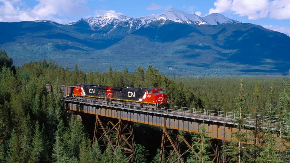 A photograph of a train crossing a bridge. There is mountain range behind the train, and the bridge is in the middle of a forest.