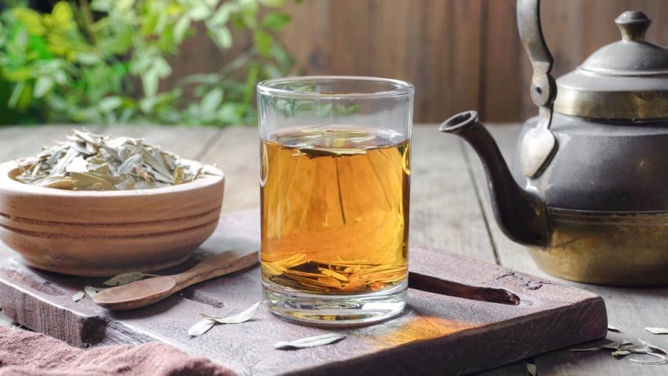 A clear glass of senna tea next to dried senna and a teapot