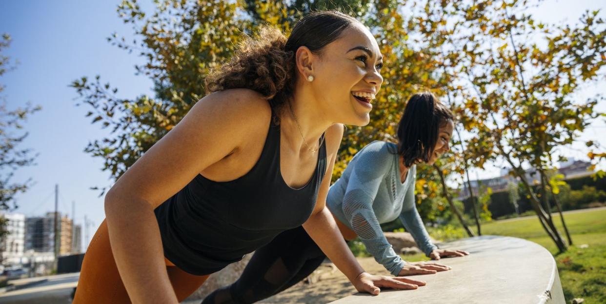 two women doing push ups on ledge outside with greenery and blue skies