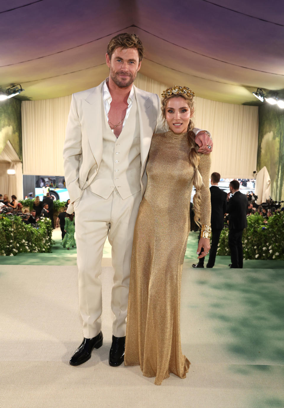Chris Hemsworth in a white suit and black shoes stands next to Elsa Pataky wearing a golden gown and tiara