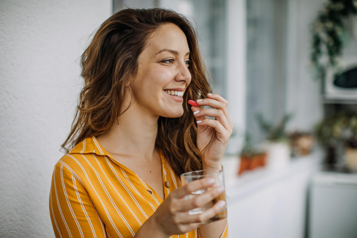 A woman holding a glass of water in her right hand closes in on popping a multivitamin with her left.