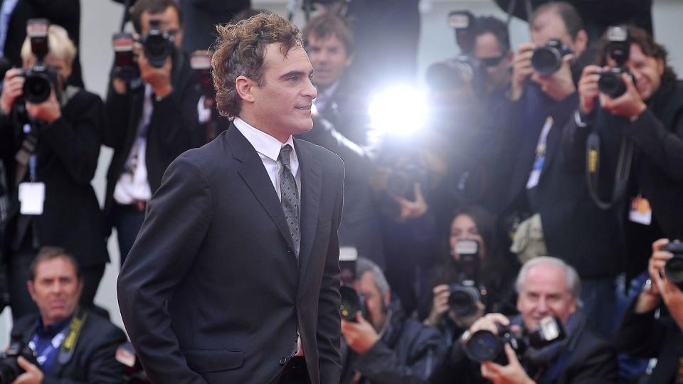 joaquin phoenix walks past a slew of photographers and smiles, he wears a black suit with a white collared shirt and gray and black patterned tie